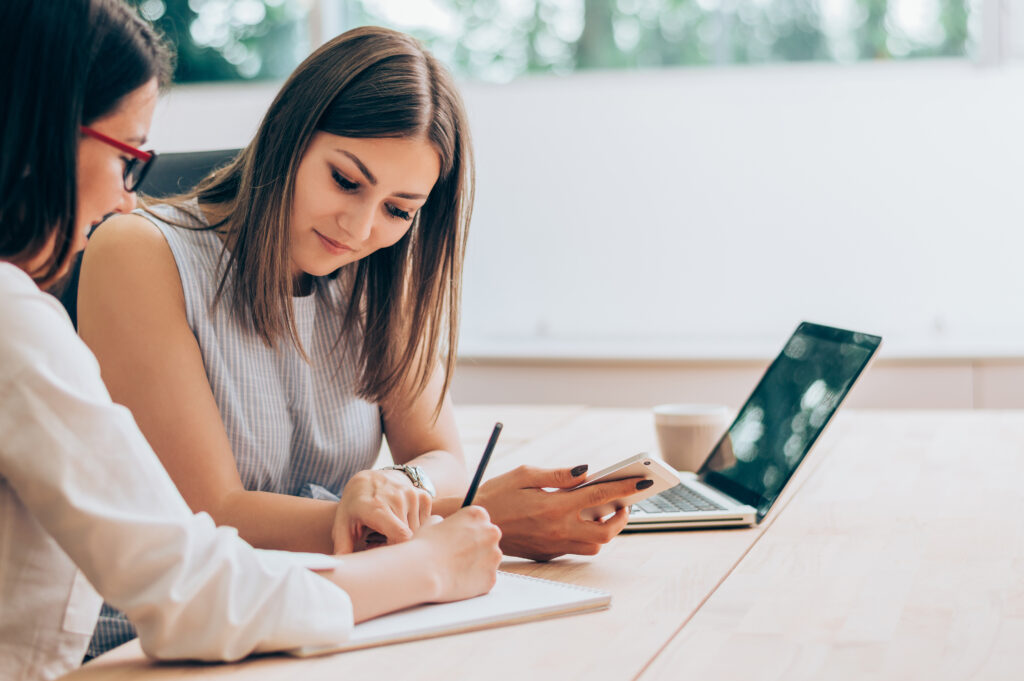 Personal growth coach working closely with a client, helping her take notes and plan goals using a notebook and smartphone in a bright and supportive environment.